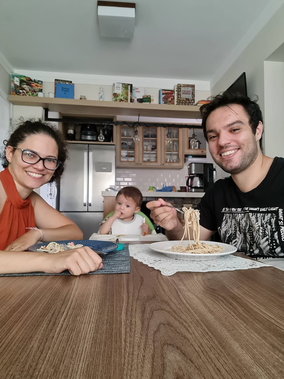 Close em Mario, um bebê de 11 meses comendo um brócolis com as mãos. Ele é branco, tem olhos claros, cabelo levemente ruivo. Seu body branco está sujo de comida, assim como seu rosto e cabelo. Apesar da boca cheia, está sorrindo, sentado no cadeirão.